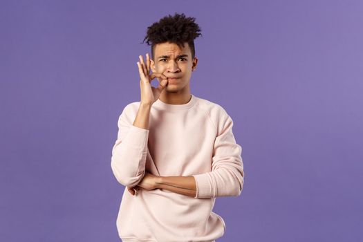 Portrait of serious-looking hispanic 25s young man promise to keep his mouth shut, secret is safe, zip his lips, lock it on key, hiding something cause he swear, standing purple background.