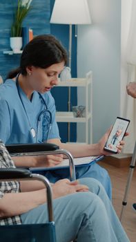 Retired woman and nurse talking to doctor on video call for telemedicine and telehealth in nursing home. Medical assistant holding smartphone, helping disabled patient in weelchair