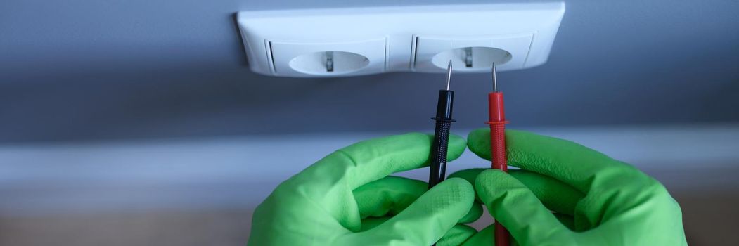 Craftsman in rubber gloves holding tester near electrical outlet at home closeup. Safety when working with electricity concept