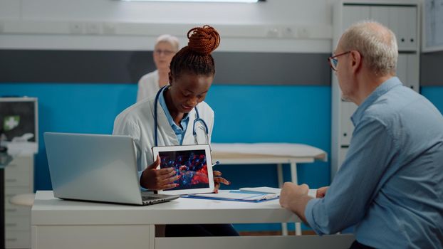 Black doctor holding virus animation on tablet showing disease cells to elder patient at desk. African american medic explaining coronavirus bacteria using modern technology on device.