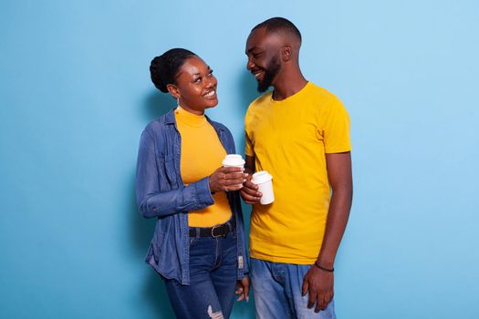 Affectionate people in love looking at each other in studio, holding cup of coffee in hand. Girlfriend and boyfriend flirting and smiling, standing over blue background and enjoying drink.