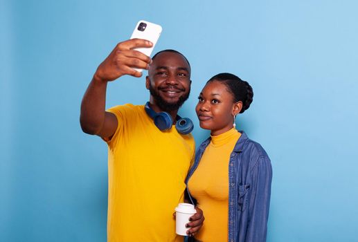 Happy couple taking selfies on smartphone in studio, making memories together. Modern boyfriend and girlfriend taking pictures on mobile phone, embracing and using technology over background.