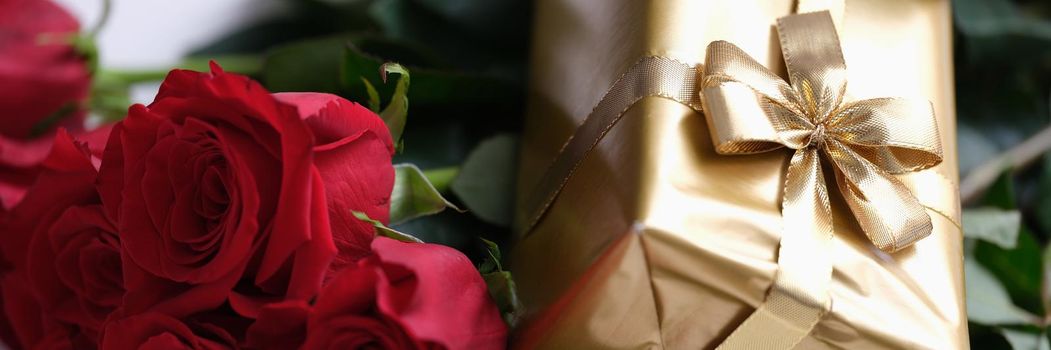 Gift in gold package with bow lying on bouquet of red roses closeup. Valentine Day concept