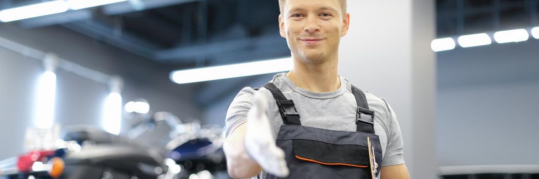 Young smiling master man in uniform stretches his arms forward in car service. High-quality service of motorcycles and cars concept