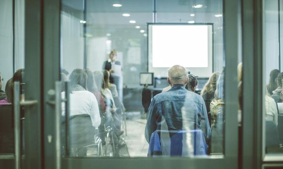 Business and entrepreneurship symposium. Female speaker giving a talk at business meeting. Audience in conference hall. Rear view of unrecognized participant in audience. Copy space on whitescreen.