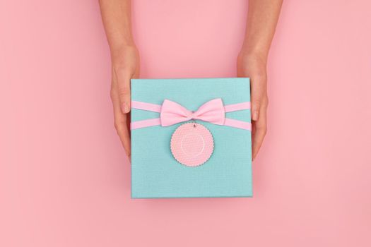 Hands give turquoise gift box with a card and a bow on isolated pastel pink background top view