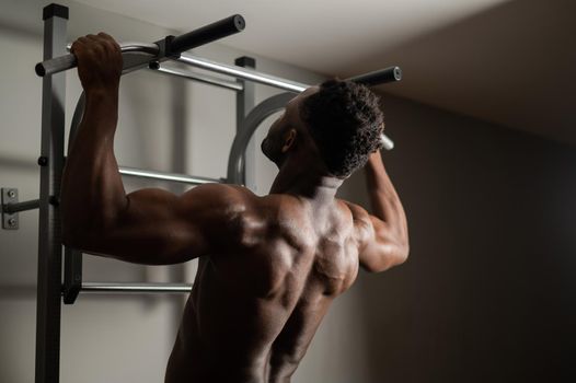 African american man with naked torso pulls up on horizontal bar in gym