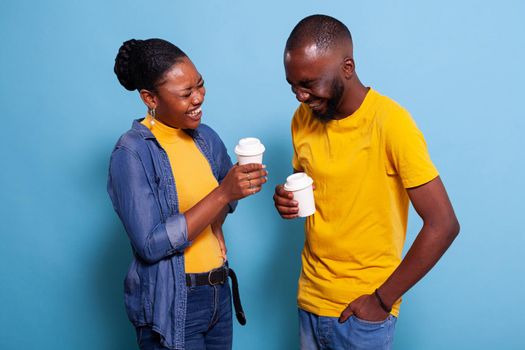 Young people laughing together and having fun in studio. Cheerful couple in love posing in front of camera and holding cup of coffee. Man and woman in relationship smiling over background.