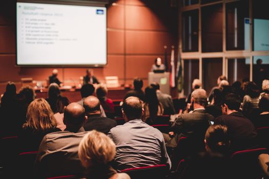 Business and entrepreneurship symposium. Speaker giving a talk at business meeting. Audience in the conference hall. Rear view of unrecognized participant in audience.