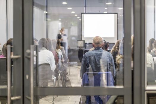 Business and entrepreneurship symposium. Female speaker giving a talk at business meeting. Audience in conference hall. Rear view of unrecognized participant in audience. Copy space on whitescreen.