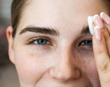 The master processes the eyebrows of a Caucasian woman with a cotton pad before laminating the eyebrows