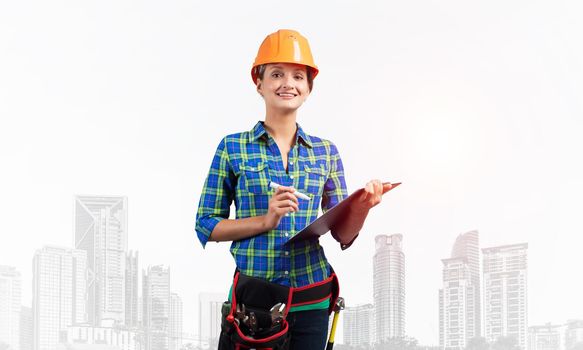 Young engineer in hardhat standing in front with clipboard checklist. Happy architect in checkered blue shirt making report on foggy metropolis background. Architecture and construction company.