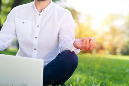 Businessman using laptop computer on green grass in park. Close up man hand in lotus pose. Man in casual wear relaxing with computer outdoors at sunny summer day. Freelance and blogging concept.