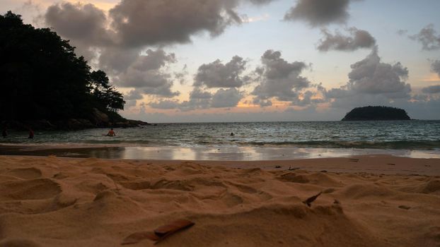 View of the beach at sunset, sea and clouds. Large clouds float across the sky. A lonely island can be seen in the distance. People relax, swim, take pictures and enjoy the sunset. Kata Beach, Phuket