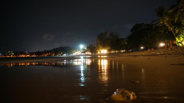 Night beach with a view of the clubs. Bright lights are burning reflecting in the sea. Fine wet sand. There is a transparent jellyfish. Palm trees grow. People are resting. Kata Beach, Phuket