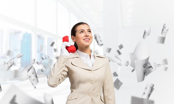 Smiling young woman holding retro red phone in office with flying paper documents. Call center operator in business suit with telephone. Hotline telemarketing. Business assistance and consultation.