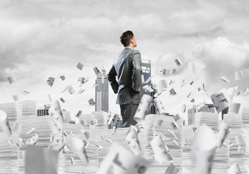 Confident businessman in suit standing on pile of documents among flying papers with cityscape on background. Mixed media.