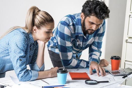 Young man and woman together working at design project. Creative teamwork at workspace with construction blueprint and color swatches. People standing near desk and discussing in architecture studio