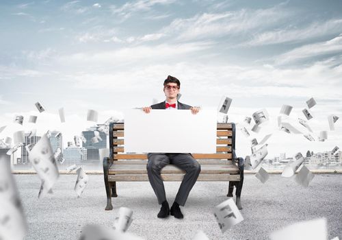 young businessman sits on a bench with an empty banner. around in the chaos flying documents