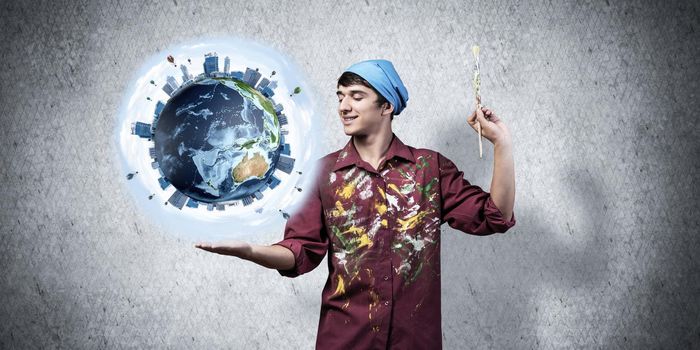 Young artist holding earth globe on hand. Male painter in dirty shirt and bandana with paintbrush on grey wall background. Creative hobby and artistic occupation. World ecology and environment concept