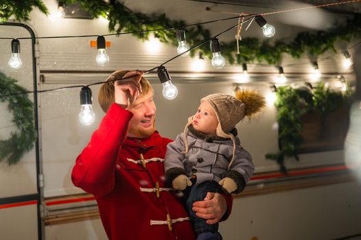 Caucasian red-haired man with a boy in his arms at the mobile home. Father and son celebrate Christmas on a trip