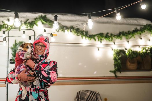 Caucasian woman holding Jack Russell Terrier dog by the trailer. Christmas van decorated.