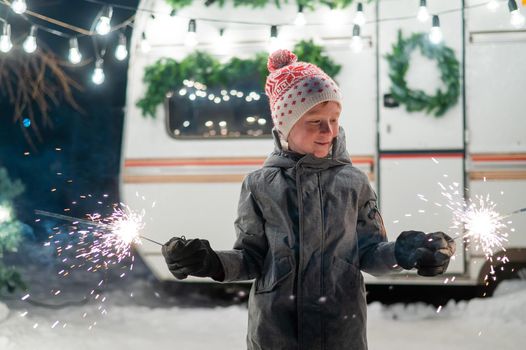Caucasian red-haired boy holding sparklers by the trailer. Schoolboy celebrates Christmas on a trip
