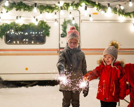 Caucasian red-haired boys hold sparklers by the trailer. Two brothers are celebrating Christmas on a trip