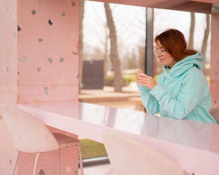 Caucasian red-haired woman eats ice cream with a plastic spoon while sitting alone in a cafe