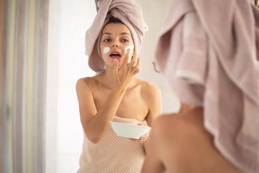 A young girl in a towel on her head and body stands near the mirror in the bathroom and applies a clay mask to her face, the woman takes care of her health and beauty.