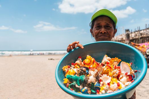 Elderly Latina street vendor on the beach displaying a bowl full of handicrafts made from painted shells. Concept of self-employment in Latin America.