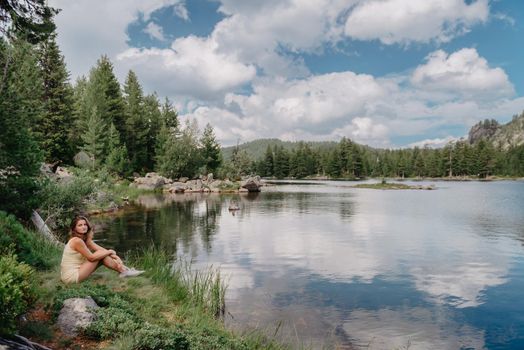 Tourist girl enjoys the magical view of the lake, coniferous forest and magical view sitting on big stone on the shore of a turquoise lake in the mountains. Hiking in the Natural Park. Black lake.
