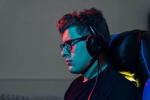 Delay of a man Young man with short blond hair, wearing glasses and headphones, dressed in black shirt, addicted to online video games, playing on a computer with headphones. Young gamer illuminated with blue and red coloured lights. Dim light from computer monitor, dark room, coloured lights. Horizontal.