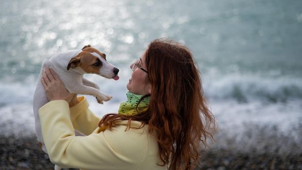 Caucasian red-haired woman holding a dog jack russell terrier on the seashore