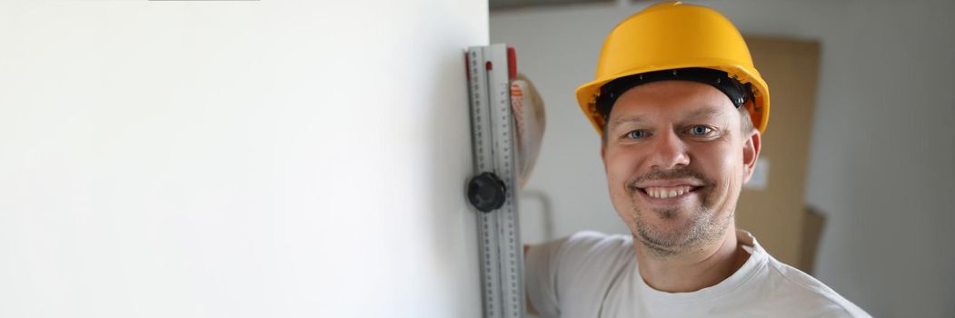 Portrait of cheerful male worker wearing protective yellow helmet checking level. Renovation in apartment. Professional carpenter. Accurate measurement and construction site concept