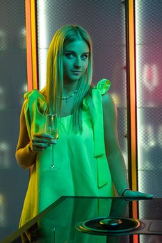 Stylish long-haired blonde girl in yellow smart dress standing with glass of sparkling wine in dimmed colored lighting of kitchen near modern induction hob. Home celebration concept