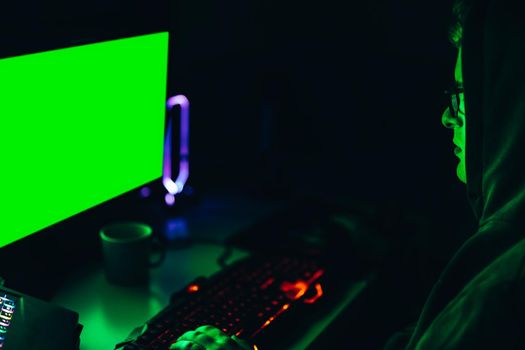 silhouette of a dangerous hooded hacker, dressed in black clothing, using computer to organise massive data breach attacks on public servers. illumination by computer monitor, green screen. Dim light from computer monitor, dark room, Horizontal.