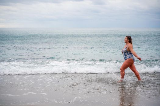 A plump woman in a bathing suit enters the water during the surf. Alone on the beach, Gray sky in the clouds, swimming in winter