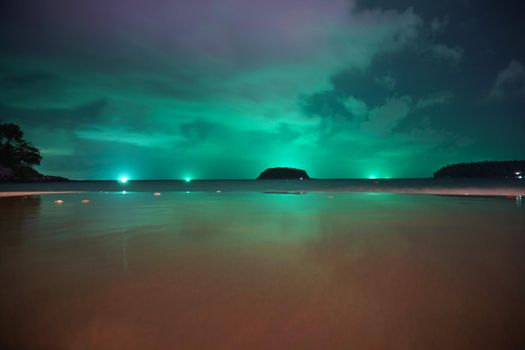 Unusual green illumination of the sky over the island. Clouds of green-purple color are reflected in the ocean, in the water and on wet sand. In distance there is an island, palm trees and lanterns