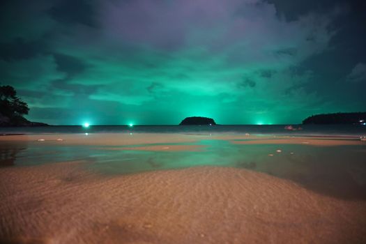 Unusual green illumination of the sky over the island. Clouds of green-purple color are reflected in the ocean, in the water and on wet sand. In distance there is an island, palm trees and lanterns