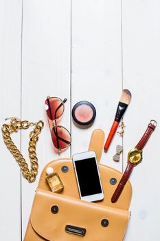 Flat lay, top view, mock up cosmetics and women's accessories fell out of the beige handbag on white background. Telephone, watches, sunglasses, perfumes