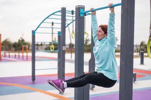 Caucasian woman in a mint sweatshirt hangs on a horizontal bar and does an exercise of the abdominal muscles on the sports ground outdoors