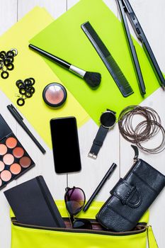 Flat lay, top view, mock up cosmetics and women's accessories fell out of the green handbag on white background. Phone, watch, pen, purse, curling