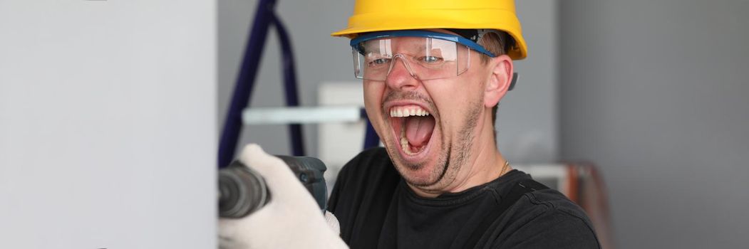 Portrait of happy worker making funny face and drill wall in room. Man in protective yellow helmet and glasses. Construction site and renovation in apartment concept