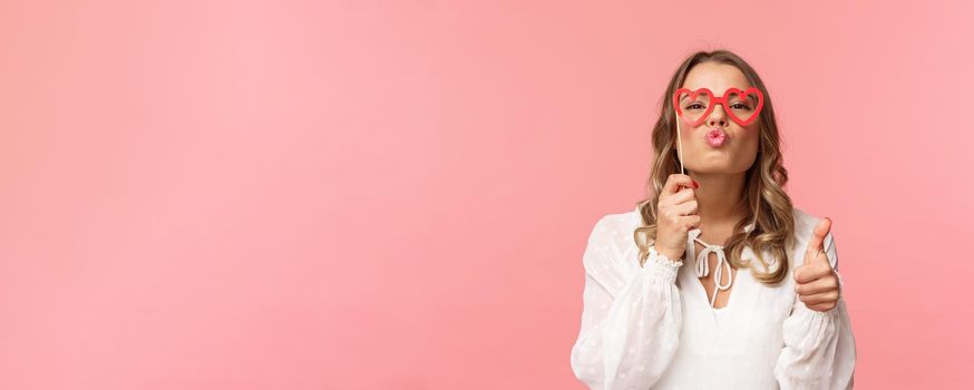 Spring, happiness and celebration concept. Close-up portrait sassy and coquettish attractive blond girl in white dress, holding heart-shaped glasses on eyes, folding lips in kiss and show thumbs-up.