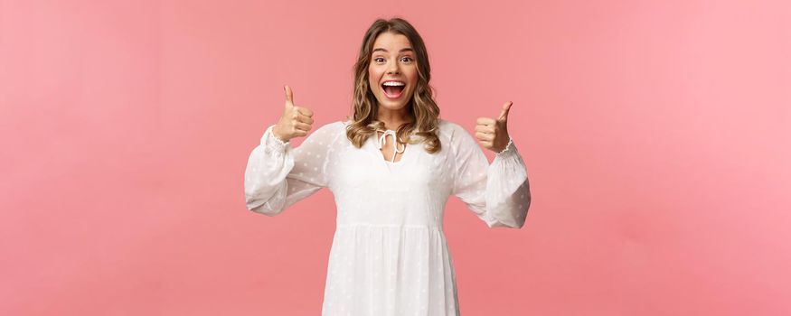 Portrait of excited feminine blond girl in white dress, show thumbs-up in approval, like being pleased, nod agreement, smiling fascinated, leave positive review, recommend product, pink background.