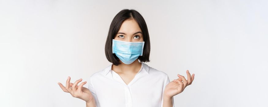 Annoyed asian woman in medical face mask, shrugging and looking up with bothered face expression, standing over white background.