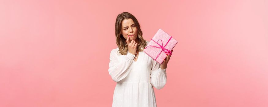 Holidays, celebration and women concept. Portrait of thoughtful curious blond girl thinking whats inside gift box, look pondering say hmm as making assumpion, standing pink background.