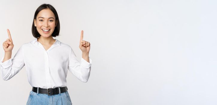 Image of smiling confident businesswoman, asian lady pointing fingers up, showing banner or sale info, standing over white background.