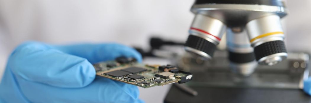 Engineer in glove holds microcircuit against background of microscope. Integrated circuit design process concept